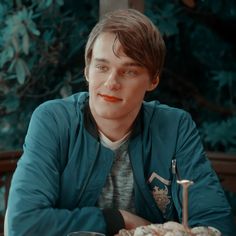 a young man sitting at a table with food in front of him and looking into the camera