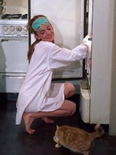 a woman in white dress kneeling next to an open refrigerator with a cat on the floor