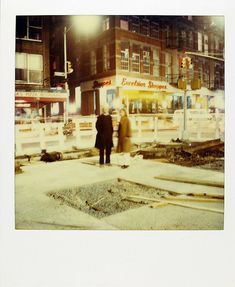 two people standing in the middle of a street at night with traffic lights and buildings behind them