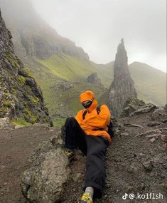 a man in an orange jacket sitting on top of a rocky hill next to mountains