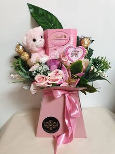 a pink teddy bear sitting on top of a box filled with flowers and chocolates