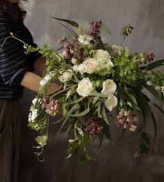 a woman holding a bouquet of flowers in front of a gray wall with greenery