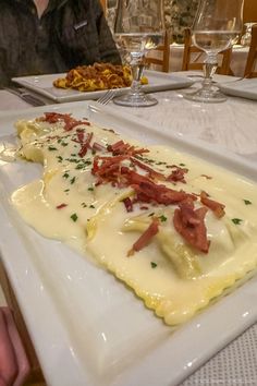 a white plate topped with ravioli covered in cheese and bacon next to wine glasses