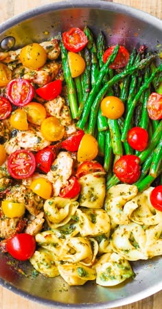 a pan filled with pasta, tomatoes and asparagus on top of a wooden table