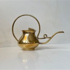 a gold colored metal tea pot with a long handle on the side, sitting on a white surface