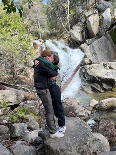 two people hugging each other on top of a rock in front of a river and waterfall