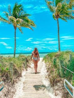 a woman walking down a wooden path between two palm trees on the beach with blue skies in the background