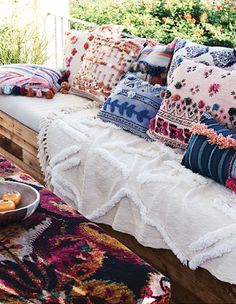 a wooden bench covered in lots of pillows on top of a rug next to a bowl of fruit