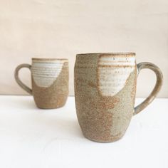 two coffee mugs sitting next to each other on a white counter top, one is brown and the other is beige