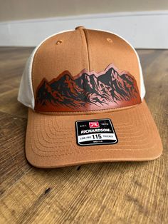 Celebrate the rugged beauty of the mountains with this exquisite Richardson 115 hat featuring the captivating mountain range leather patch. Whether you're exploring nature trails, summiting peaks, or simply enjoying the view, this hat is the perfect companion for your outdoor escapades. This Richardson 115 Caramel/Birch hat featuring a striking leather patch depicting a mountain range has been meticulously matched by our color professionals. This hat and patch combination is perfect for hikers, Custom Leather Hat Patch, Brown Western Style Baseball Cap For Outdoor, Adjustable Western Baseball Cap For Outdoor, Western Style Adjustable Baseball Cap For Outdoor, Brown Western Snapback Hat For Outdoor, Country Style Brown Baseball Cap For Outdoor, Country Style Baseball Cap For Outdoor, Rugged Brown Hat For Travel, Rugged Brown Travel Hat