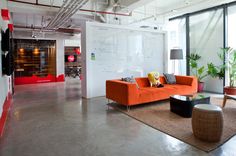 an orange couch and two chairs in a room with large windows, rugs and potted plants on the floor