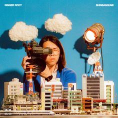 a woman holding a camera in front of a model city with clouds above her head