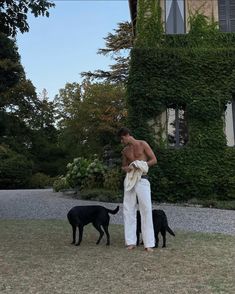 a shirtless man standing next to two black dogs in front of a large house