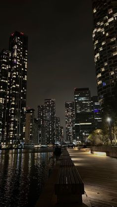 the city skyline is lit up at night with lights reflecting in the water and benches along the waterfront