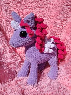 a purple crocheted pony sitting on top of a pink fuzzy blanket with flowers in its hair