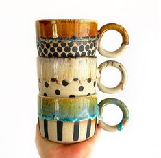 a hand holding a stack of four coffee mugs in different colors and designs on them