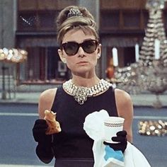 a woman in black dress and sunglasses holding a white bag while standing next to a building