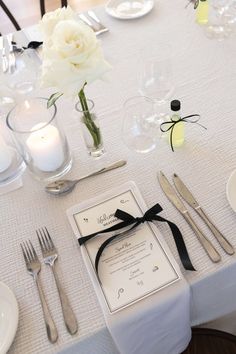 the table is set with silverware and white roses in vases, candles, and menu cards