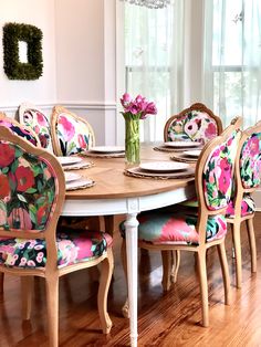 a dining room table with colorful chairs and flowers on the chair backrests, in front of a window