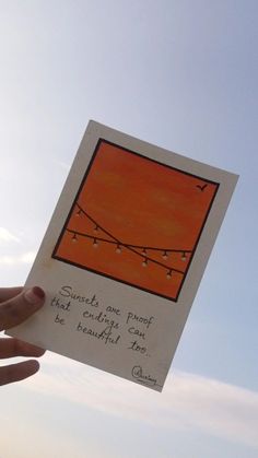 a hand holding up a piece of paper with string lights on it and an orange sky in the background