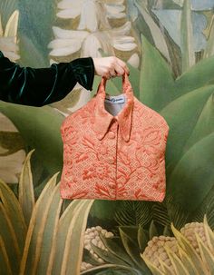 a woman holding an orange purse in front of a wall with plants and flowers on it