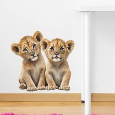 two lion cubs sitting next to each other in front of a white wall and pink rug