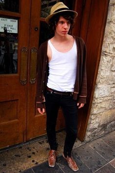 a young man standing in front of a wooden door wearing a hat and black pants