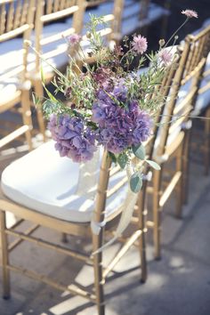 purple flowers are in a vase on the back of a chair at an outdoor ceremony