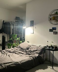 an unmade bed in a small room with plants on the table and clothes hanging up