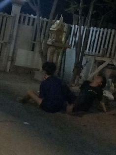 two people sitting on the ground in front of a white fence at night with one person holding a cell phone