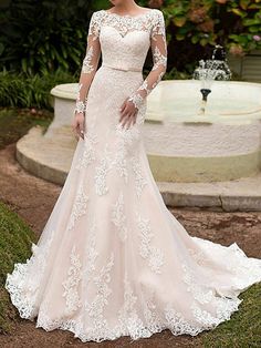 a woman wearing a wedding dress with long sleeves and lace on the top, standing in front of a fountain