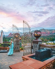 a woman in a blue dress standing next to an iron gate and fountain at sunset