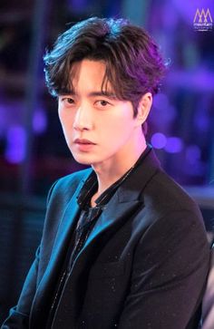 the young man is wearing a black suit and looking off into the distance with purple lights behind him