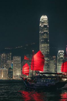 a boat with red sails sailing in the water near tall buildings at night, hong kong