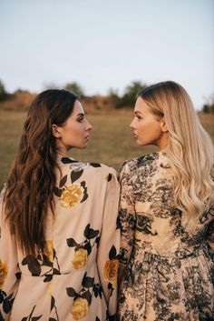 two young women standing next to each other in front of a field with grass and trees