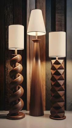 Three wooden lamps with unique designs a spiral base, a tall slender base, and a faceted geometric base, all with white cylindrical shades, are displayed against a dark wooden background. #unique #nightlamp #design #ideas Large Wooden Lamps, Wood Column Floor Lamp, Tall Timber Lamp, Cnc Lamp Wood Lighting Design, Wooden Lamp Base, Modern Bungalow Exterior, Wooden Lamps, Kitchen Tools Design, Steampunk Lamp Wood