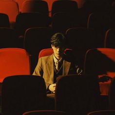 a man in a suit and tie sitting in an empty auditorium with rows of red chairs