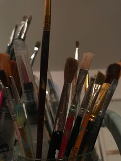 many brushes are in a clear cup on the counter next to some pens and pencils