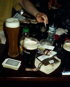 A table in a dimly lit pub filled with glasses of beer, ashtrays a deck of cards, lighters and tabac . A hand reaches to ash a cigarette above the ashtray. Club Film Photography, 60s London Aesthetic, Beer Glass Aesthetic, Pub Instagram Ideas, Pub Owner Aesthetic, 80s British Aesthetic, Pub Vibes Aesthetic, Pub Drinks, Making Drinks Aesthetic