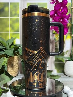 a black and gold travel mug sitting on top of a plate next to purple flowers