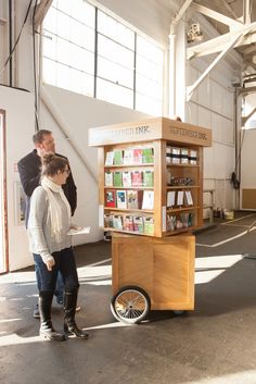 a man and woman standing in front of a cart with books on it while another person stands next to the cart