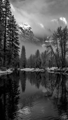 a black and white photo of a lake surrounded by trees with snow covered mountains in the background