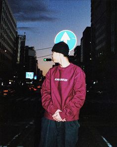 a young man standing in the middle of a city at night wearing a maroon sweatshirt and black pants