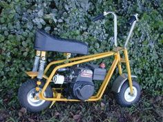 a small yellow motorcycle parked in front of some bushes and grass with no wheels on it