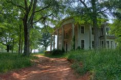 an old house in the middle of a dirt road surrounded by trees and greenery