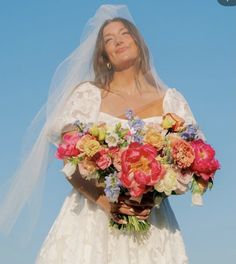 a woman wearing a wedding dress holding a bouquet of flowers in front of a blue sky