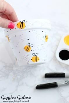 a hand holding a cup with bees on it next to some markers and paintbrushes