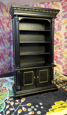 an antique black bookcase with gold trimmings on the front and sides, sitting on a floral patterned tablecloth