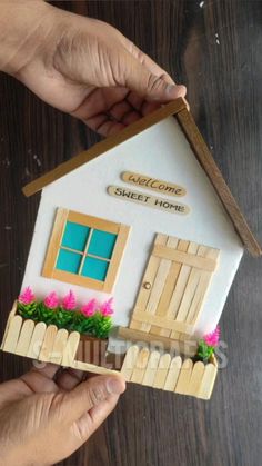 two hands holding a small house made out of popsicle sticks and some fake flowers