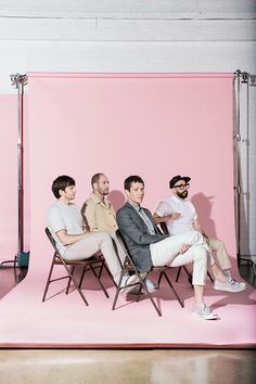 three men sitting on folding chairs in front of a pink wall, with one man looking at the camera